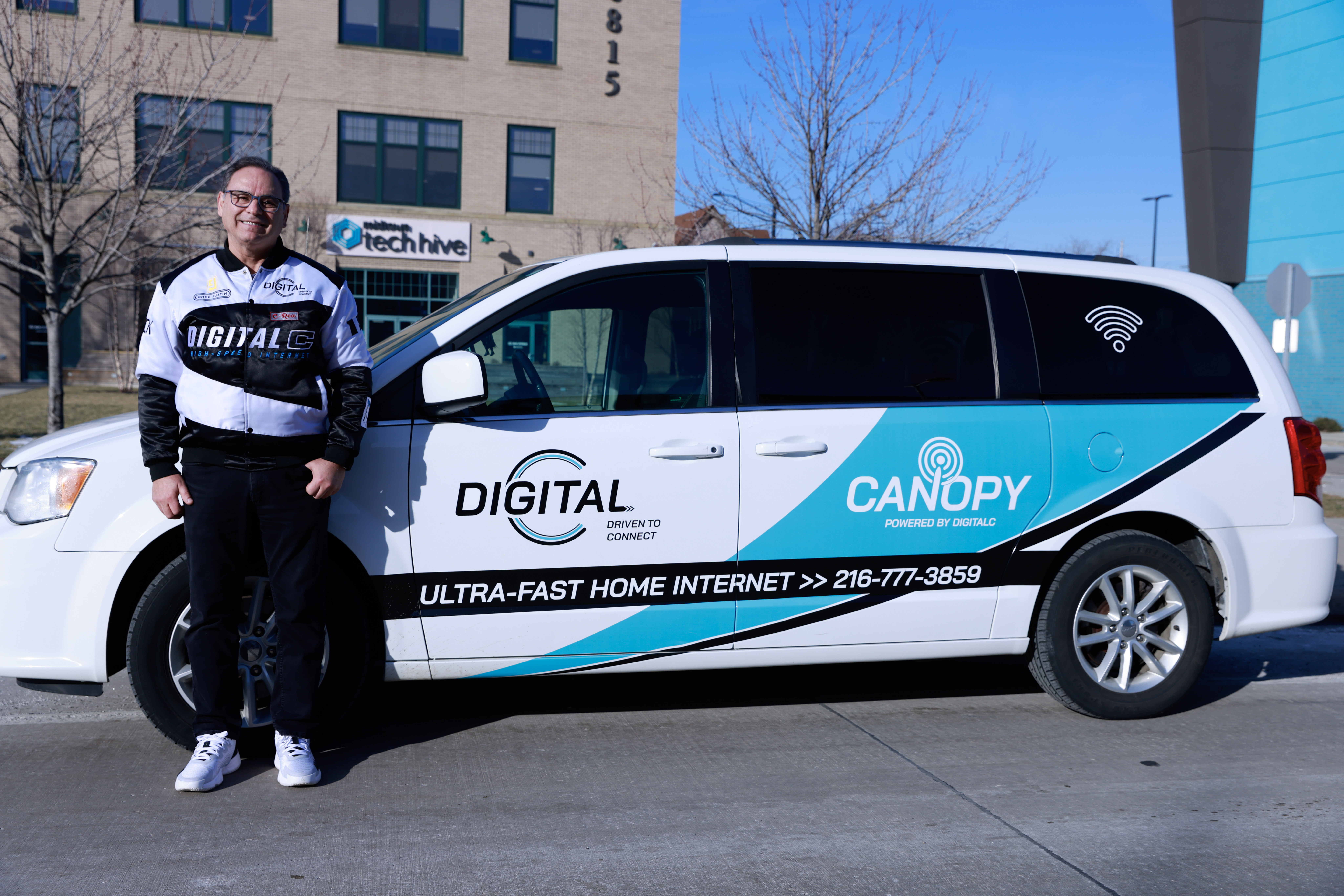 Man wearing a DigitalC bomber jacket stands in front of a van wrapped in DigitalC branding, parked outside the MidTown Tech Hive headquarters.